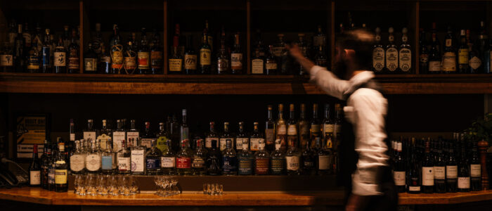 A bartender moves behind a well-stocked bar, reaching for a bottle on the top shelf. The background features an extensive liquor selection and decorative plates on the wall.