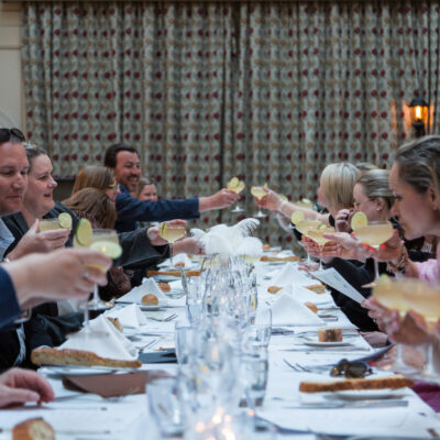 A lively banquet scene where a long table of guests raises their glasses for a toast. The table is set with white linens, bread, and drinks garnished with lime slices, while guests smile and engage with one another.