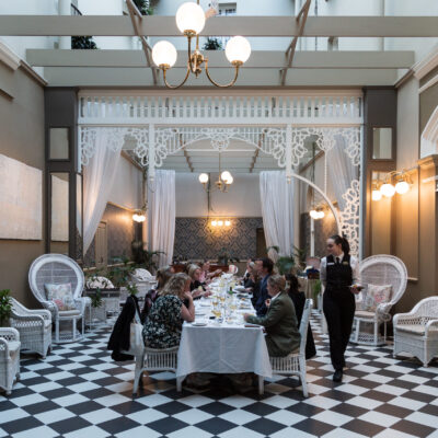 A beautifully designed conservatory with a black-and-white checkered floor and white wicker furniture. A long dining table is set for an elegant meal, with guests seated and a waiter in a black uniform serving drinks.