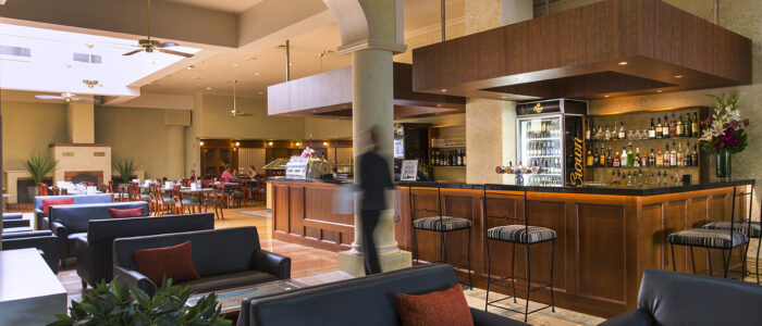 A hotel bar. Numerous black sofas with coffee tables in front of them. A staff member in black walks out of focus in the center of the image. Behind her is modern looking bar, further on is a restaurant area where two people are sitting at a table