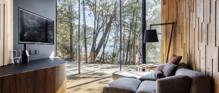 A cozy modern living room with large floor-to-ceiling glass windows offering a view of tall trees and a distant body of water. The room features warm wooden paneling, a plush gray armchair with cushions, a sleek wall-mounted TV, and a black standing lamp. A wooden cabinet holds a bottle of wine and two glasses.