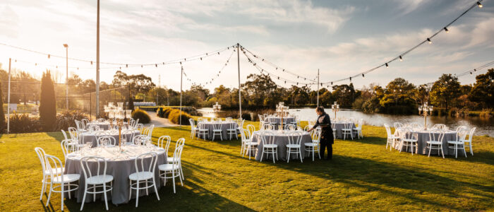 A lawn area next to a lake. Multiple round tables set on lawn with white chairs. Waiter is setting one of the tables.