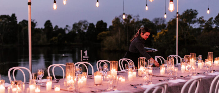 An outdoor dining setup at dusk featuring a long table adorned with glowing candles, glassware, and white chairs. String lights hang above, and a server arranges table settings in front of a serene lakeside view with trees in the background.