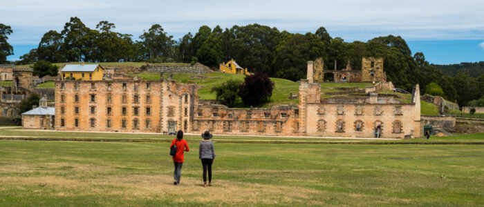 Two people walking on a grass field towards old sandstone brick buildings. Behind this building is a hill with more stone buildings scattered around the hill