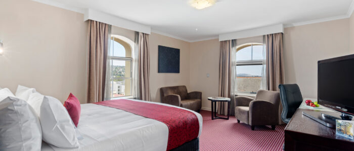 A cozy bedroom with stripped red and white carpets. Neatly made double bed, two chairs in the corner with a coffee table and a tv to the right of the image on a dark hard wood cabinet.