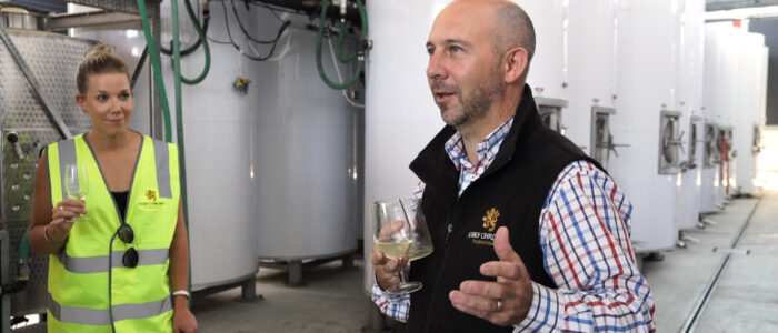 A man and a woman stand inside a winery near large stainless steel tanks. The man, wearing a black vest and holding a wine glass, is speaking while gesturing with his hand. The woman, in a bright yellow safety vest, also holds a wine glass and listens attentively.