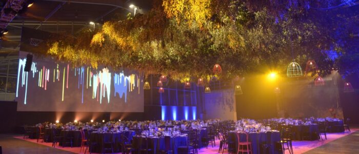 Indoor event venue, plants and lights hanging from the center of the room. Concrete floor with many round set tables. Large screen on the back wall with art displayed - looks like paint dripping