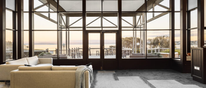 Interior of a contemporary lounge with a natural aesthetic, showcasing wooden features and spectacular views of Oyster Bay. White couch to the left. Large glass windows.