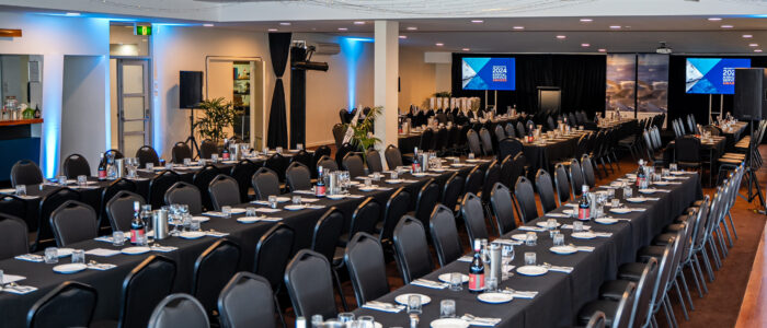 Boathouse-Conference_2 A banquet hall with long tables draped in black tablecloths, set with plates, glassware, and cutlery. Rows of black chairs surround the tables. The back wall has to screens displaying "2024 Annual Service Awards". The ceiling is adorned with string lights.