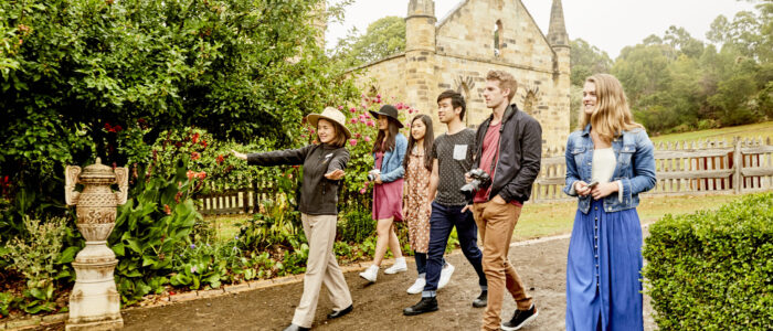 Group of five people being led by a woman in a black jacket along a path surrounded by trees, plants and garden ornaments. Behind them is a old stone chapel surrounded by a wooden pickeered fence.