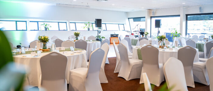 Boathouse-Conference_3 A decorated event space with round tables covered in white tablecloths and green table runners. The tables are elegantly set with glassware, cutlery, and floral centerpieces. White chairs with covers surround the tables, and a small stage with musical instruments is visible in the background.