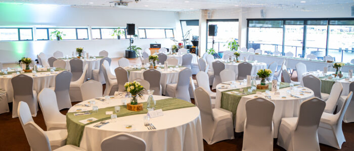 Boathouse-Conference_4 A decorated event space with round tables covered in white tablecloths and green table runners. The tables are elegantly set with glassware, cutlery, and floral centerpieces. White chairs with covers surround the tables, and a small stage with musical instruments is visible in the background.