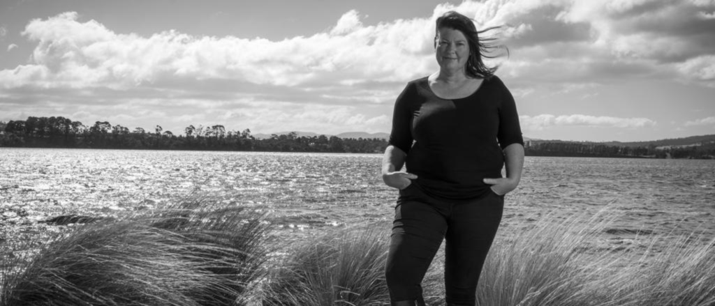 The image shows Natalie Fryar standing outdoors near a body of water with tall grass around. She is wearing dark clothing and boots. The background includes water, distant trees, and a partly cloudy sky. The image is in black and white.