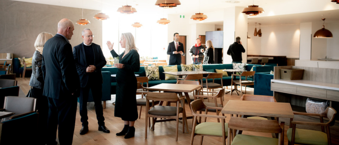 A group of people standing and conversing in a modern, well-lit dining area or cafeteria. The space is furnished with wooden tables and chairs, some of which have green cushions. There are several pendant lights hanging from the ceiling, and the overall decor appears contemporary. In the background, more people can be seen either standing or walking around.