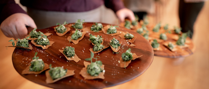 A person holding two wooden trays filled with appetizers. The appetizers appear to be small portions of a green mixture, possibly a type of salad or spread, placed on top of triangular pieces of crispy bread or crackers. The trays are being held by the person's hands, and the background is slightly blurred, indicating an indoor setting.