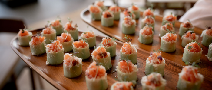 Two wooden trays filled with neatly arranged pieces of sushi. The sushi pieces appear to be wrapped in cucumber and topped with a mixture of raw fish, possibly salmon, and garnished with small green herbs.