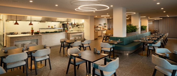 A modern, well-lit restaurant interior with a clean and minimalist design. The seating area features several tables with chairs arranged neatly. The tables are set with small plates, cups, and cutlery. There is a long counter in the background with an open kitchen area where cooking equipment and ingredients are visible. The ceiling has circular light fixtures that add to the contemporary ambiance of the space. There are also some plants placed around the restaurant, contributing to a fresh and inviting atmosphere.