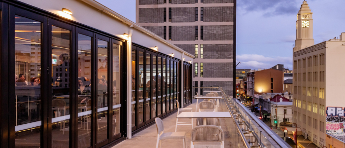 An outdoor terrace with tables and chairs along the railing, adjacent to a glass-walled indoor area where people are seated. In the background, buildings and a tall clock tower are visible, with dusk lighting and a softly colored sky.