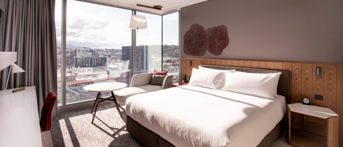 A modern hotel room with a large bed, a small round table with two chairs, a desk, and floor-to-ceiling windows offering a city view. The room features neutral tones and contemporary decor.