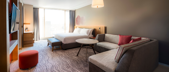 A modern hotel room with a large bed, a sofa, a round coffee table, and a flat-screen TV. The room has large windows with curtains that allow natural light to fill the space. There is also an ottoman and a desk area near the window. The decor is contemporary with neutral tones and red accents.
