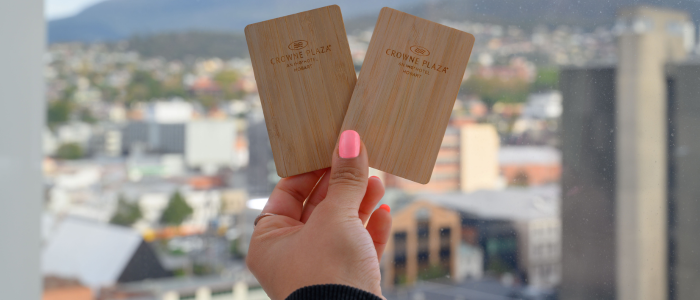 A hand holding two wooden key cards with the text "CROWNE PLAZA" and "Hobart" printed on them. The background features a cityscape with buildings and mountains in the distance.