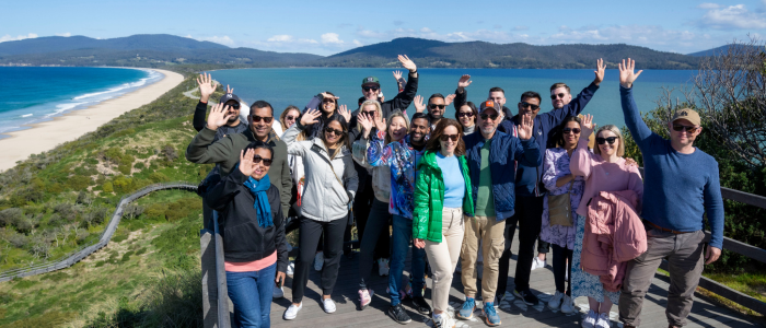 Derwent Exp (Do) 1 A group of people is standing on a wooden platform overlooking a scenic coastal landscape. The background features a long sandy beach, blue ocean waters, and distant hills under a partly cloudy sky. The individuals are waving at the camera.