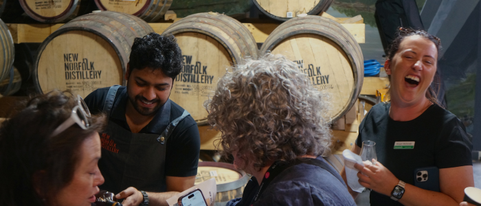Derwent Exp (Do) 2 The image shows a group of people standing in front of several wooden barrels labeled 'NEW NORFOLK DISTILLERY.' The people appear to be engaged in a tasting or tour, as one person is holding a glass, and another is holding a bottle.