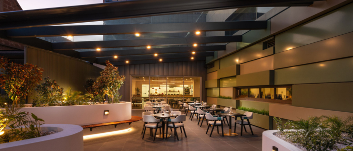 A modern outdoor dining area with several tables and chairs arranged neatly. The space is well-lit with ceiling lights, and there are plants in large planters along the sides. In the background, there is an indoor section visible through large glass windows, which appears to be part of a restaurant or café. The overall design is contemporary with clean lines and a mix of natural and artificial lighting.