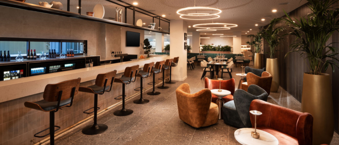 A modern, stylish bar and lounge area. There is a long bar counter with several high chairs lined up in front of it. Behind the bar, there are shelves with various bottles and glasses, as well as a small refrigerator. The ceiling features circular light fixtures that provide ambient lighting. In the foreground, there are comfortable armchairs and small tables arranged for seating. The background includes more seating areas with tables and chairs, along with some greenery adding to the decor.