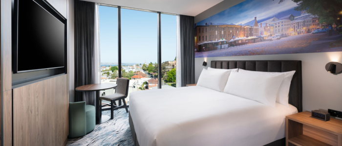 A modern hotel room with a large bed, a flat-screen TV mounted on the wall, a small round table with two chairs, and floor-to-ceiling windows offering a view of the cityscape outside. The room has contemporary decor with neutral tones and features a large mural of an outdoor scene on the wall behind the bed.