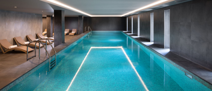 An indoor swimming pool with a modern design. The pool is rectangular, with clear blue water and tiled flooring. On the left side of the pool, there are several lounge chairs arranged in a row, each with a white towel placed on it. The walls and ceiling are finished in dark tones, giving the space a sleek and contemporary look. There are also several vertical windows on the right side of the pool area, allowing natural light to enter. The lighting is recessed into the ceiling, providing an even illumination throughout the space.