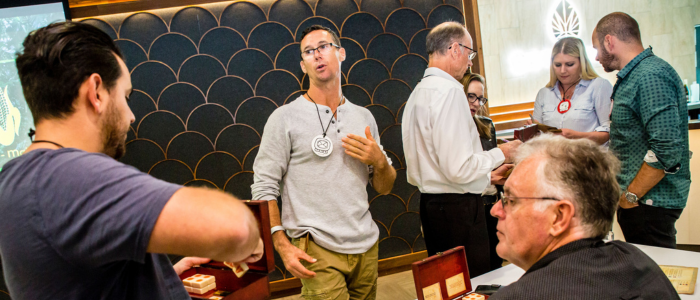 Experiential Edge 3 A group of adults interacting during a workshop in a modern indoor venue. A man at the center gestures while speaking, wearing a round badge, as others listen, handle small wooden boxes, or converse in pairs. The background features patterned wall panels and warm lighting, reinforcing a professional, collaborative event setting.