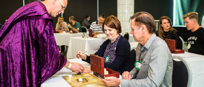 Experiential Edge 4 Two adults seated at a table participate in a hands-on workshop activity, examining a small wooden box and written materials. A facilitator wearing a purple cloak leans in to assist, holding an object over a board laid out on the table. Other participants work in the background, creating a focused, immersive atmosphere in a modern event space.
