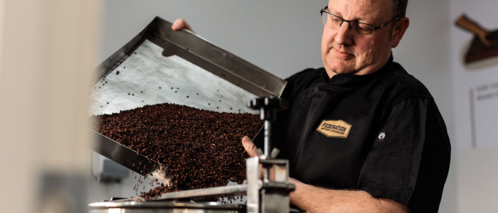 A person wearing a black apron with a name tag is pouring cocoa from a metal tray into a machine.