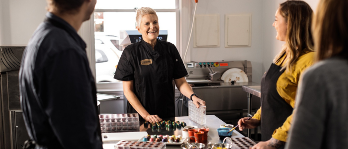 A group of people in a kitchen or culinary setting. One person, wearing a black uniform and name tag, appears to be demonstrating or explaining something to the others. Various kitchen tools and ingredients are on the counter, including molds, bowls with different contents, and what looks like chocolates or confections. The background includes typical kitchen equipment such as an oven and other appliances.