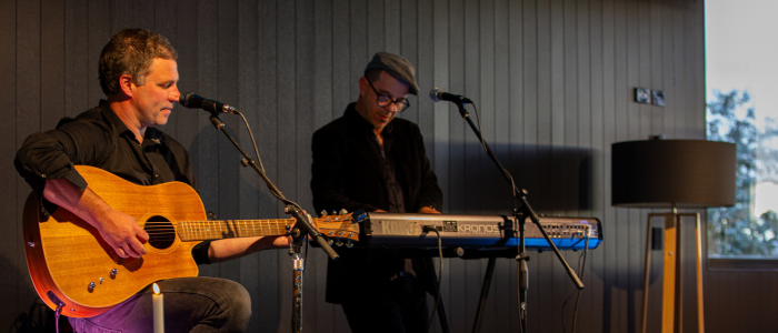 Joel Everard Music 2 2 people on stage. One playing guitar and the other playing the keyboard. Black wooden wall behind them
