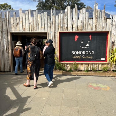 Entrance to Bonorong Wildlife Sanctuary featuring a rustic wooden facade and a large black sign with a stylized animal face and red ears, with people walking toward the doorway on a sunny day.
