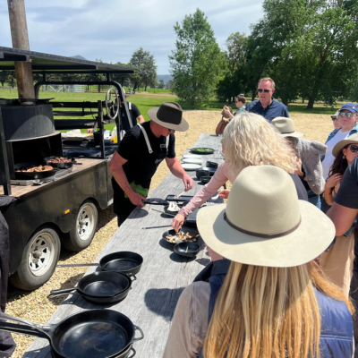 Outdoor cooking demonstration with a chef serving food from a large black barbecue trailer onto plates on a wooden table, surrounded by people wearing hats in a sunny open area with trees in the background.