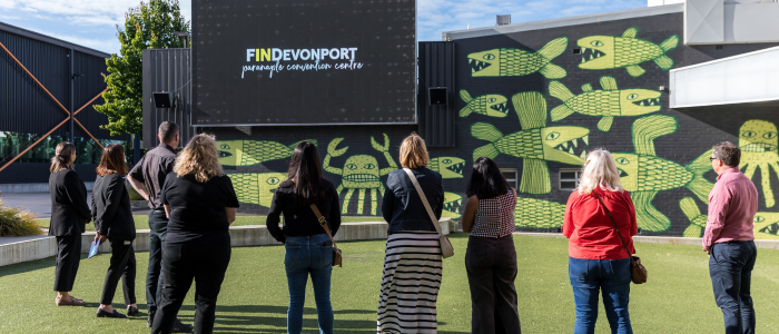 A group of people standing on a green lawn, facing a large screen mounted on a building. The screen displays the text 'FINDevonport' and 'paranaple convention centre.' The building features a mural with green fish-like creatures.