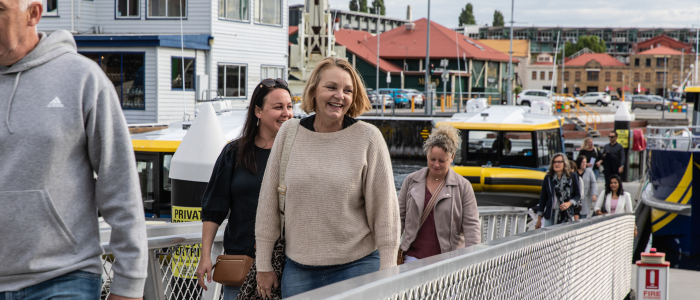 People walking along a metal gangway at a waterfront marina, with yellow and white boats docked nearby and buildings with red roofs in the background.