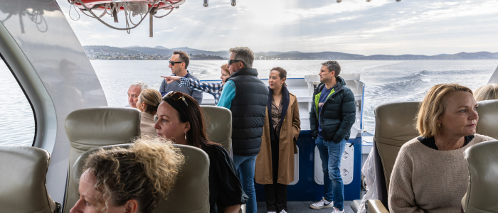 Passengers seated inside a modern tour boat with large windows, while several people stand at the rear deck looking out over calm water toward distant hills under a partly cloudy sky.