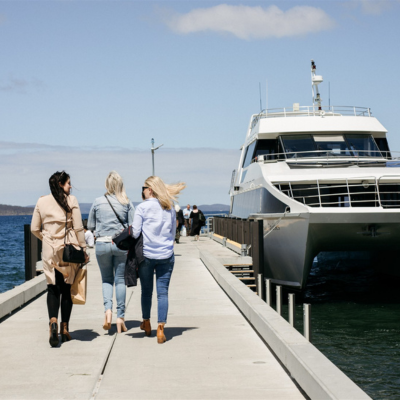 Three people walking along a concrete pier toward a large white catamaran-style boat docked at the end, with calm blue water on both sides and a clear sky above.