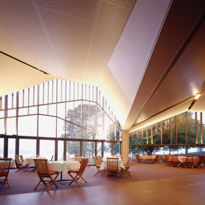 Spacious modern dining area with high angled ceilings and large floor-to-ceiling windows overlooking trees and water, featuring neatly arranged tables with white tablecloths and wooden chairs under soft natural light.