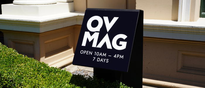 Queen Victoria Museum and Art Gallery 3.0 A black metal sign that reads "QVMAG. Open 10am - 4pm, 7 Days" This sits hanging over a green, neatly trimmed hedge with a stone building behind