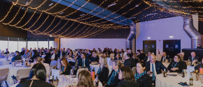 Tailrace Centre (Conference) 1 A large banquet hall filled with attendees dressed in formal attire, seated at round tables. The ceiling is adorned with warm string lights, creating an elegant atmosphere. Many guests appear engaged, looking toward an unseen speaker or presentation.