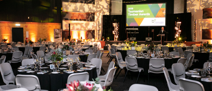 Tailrace Centre (Conference) 2 A beautifully decorated event hall set up for the 2019 Tasmanian Timber Awards. Round tables with black tablecloths, fine glassware, and floral centerpieces fill the room. A stage with a large screen displaying the event name is in the background, along with warm ambient lighting.