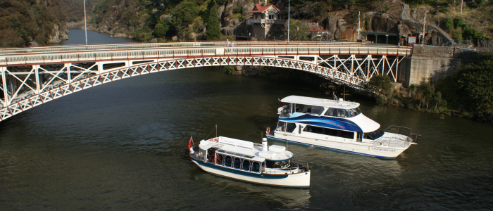 Two boats navigate a calm river beneath a white iron bridge, surrounded by a rocky gorge with lush greenery. A house sits on the hillside, overlooking the scene.