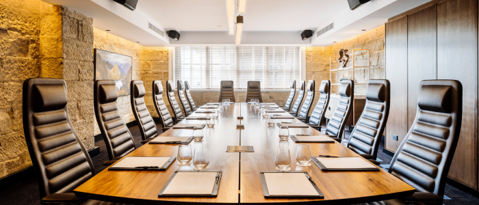 Tasman (Conference) 1 A modern conference room with a long wooden table surrounded by twelve high-back leather chairs. Each seat is set with a notepad and a glass of water. The room features stone walls, large windows with blinds, ceiling lights, mounted speakers, and a piece of artwork on one wall.