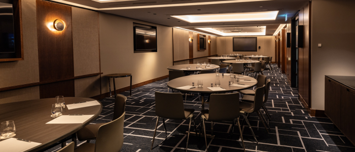 Tasman (Conference) 2 A modern conference room featuring round tables surrounded by chairs, each table set with glasses of water, notepads, and pens. The room has a patterned carpet, wood-paneled walls, recessed ceiling lighting, and several flat-screen televisions mounted on the walls.