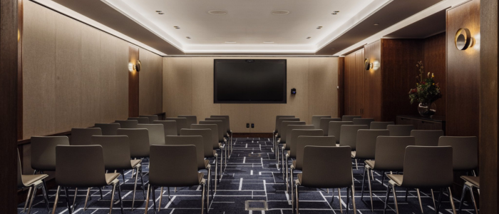 Tasman (Conference) 3 conference room with rows of gray chairs facing a large flat-screen TV mounted on the front wall. The room features beige walls, dark wood paneling, a patterned carpet, wall sconces for lighting, and a flower arrangement on a table to the right.